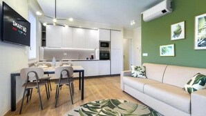 A modern kitchen with white cabinets, a dining area with chairs, a sofa, and a green wall with framed pictures. - Hostdomus - Green House (Diano Marina)