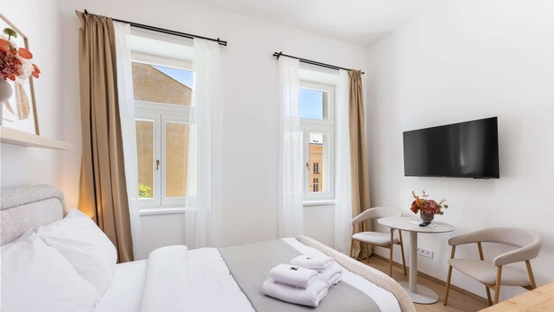 A hotel room with a bed, two windows with curtains, a TV mounted on the wall, a small round table with chairs, and a wooden desk. - Lovely studio in the center city (Hlavní město Praha)
