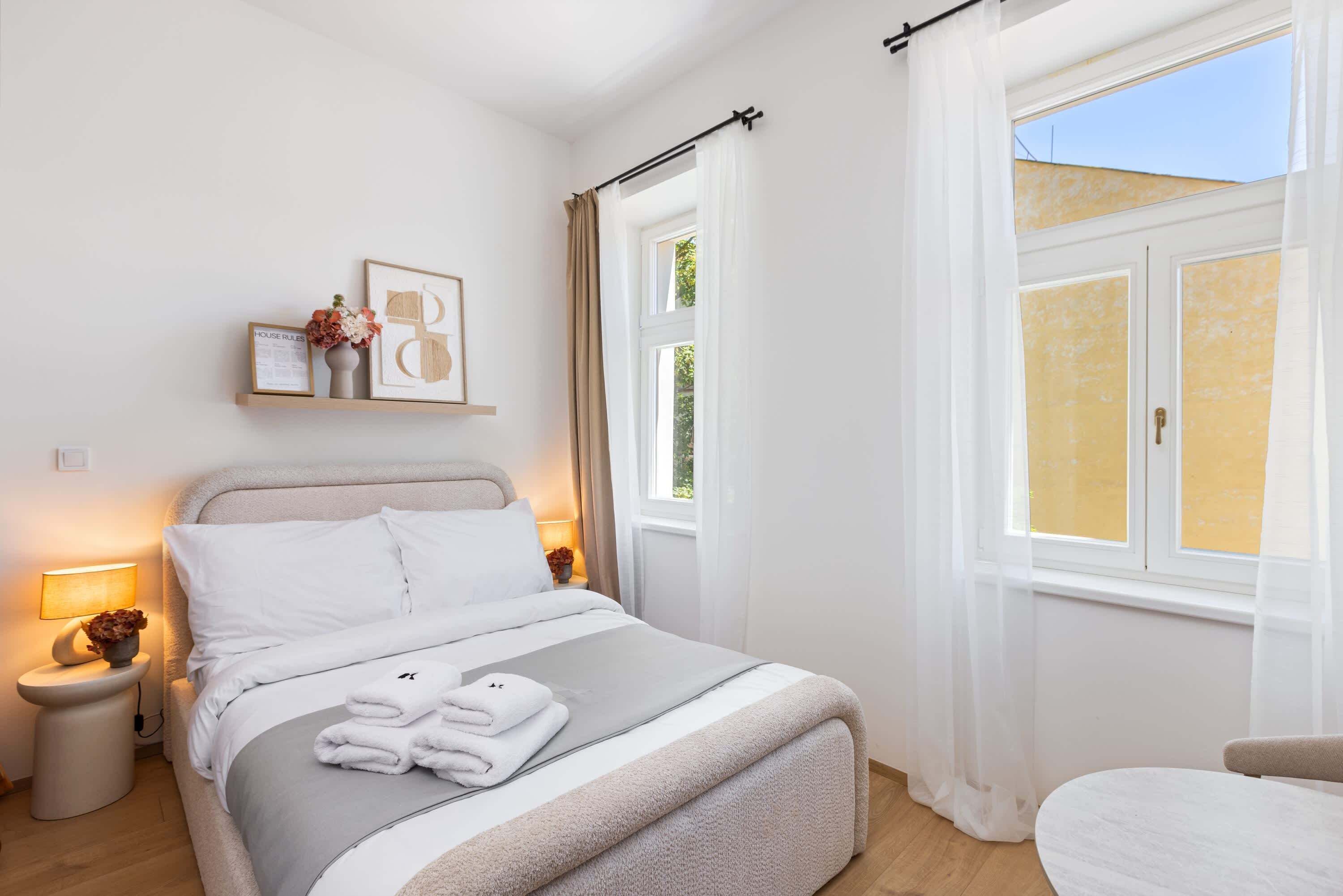 Un lit bien fait, avec une couette grise et blanche, une table de chevet avec une lampe et une fenêtre ornée de rideaux légers.