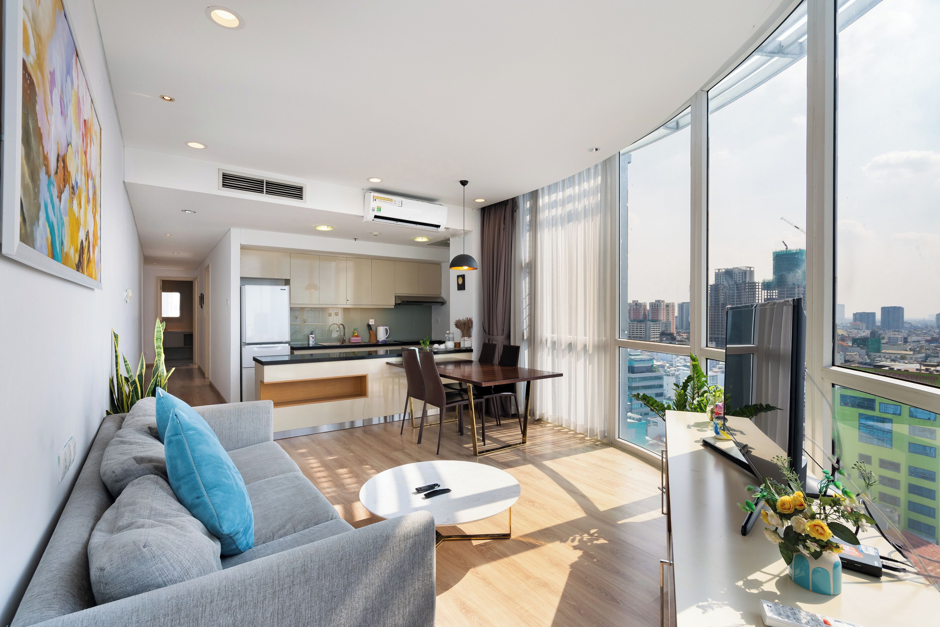 A modern apartment with a living room, dining area, and kitchen. It features large windows, a city view, and a balcony with plants.