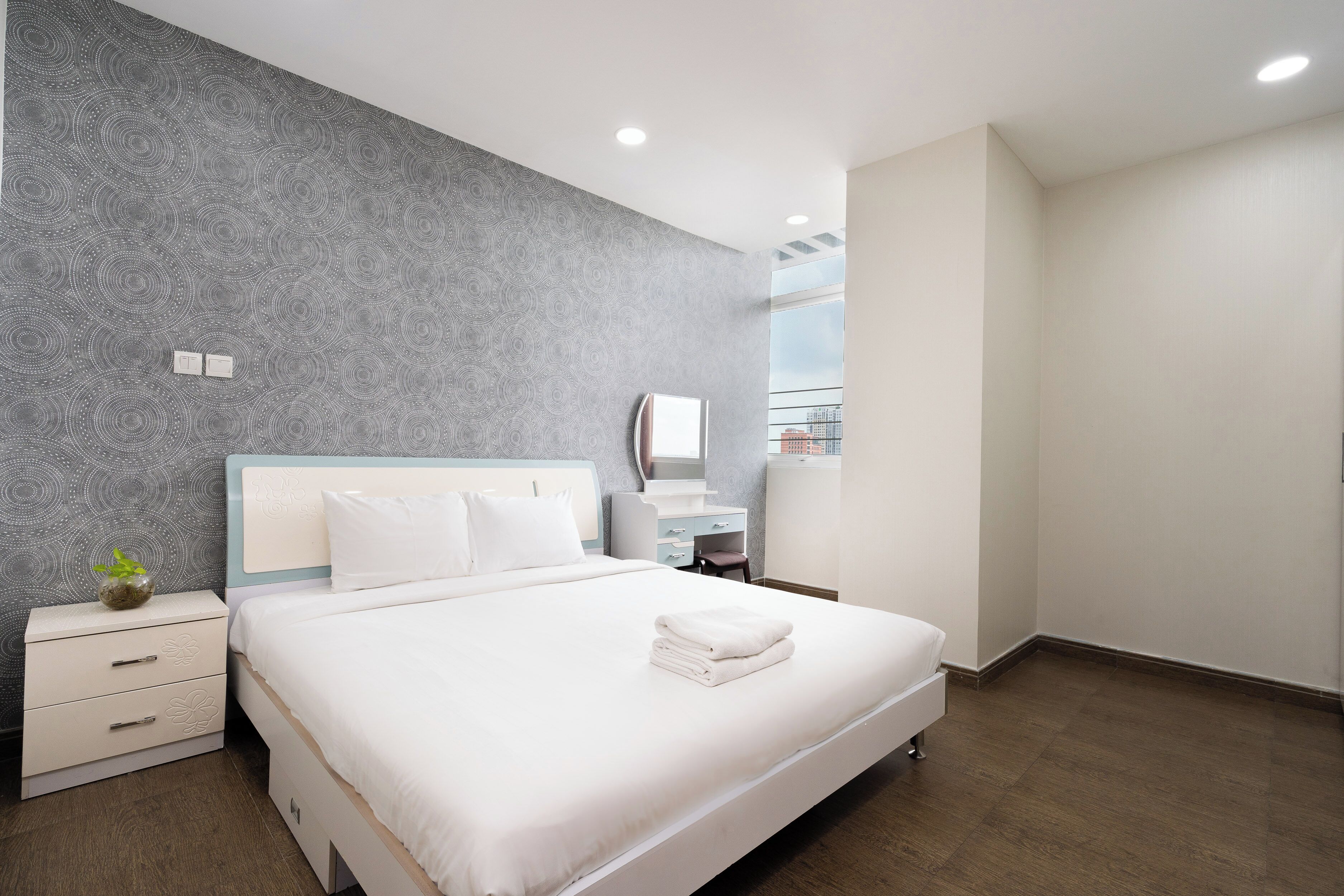 A modern hotel room with a large bed, a bedside table, a dresser, and a view of the city.