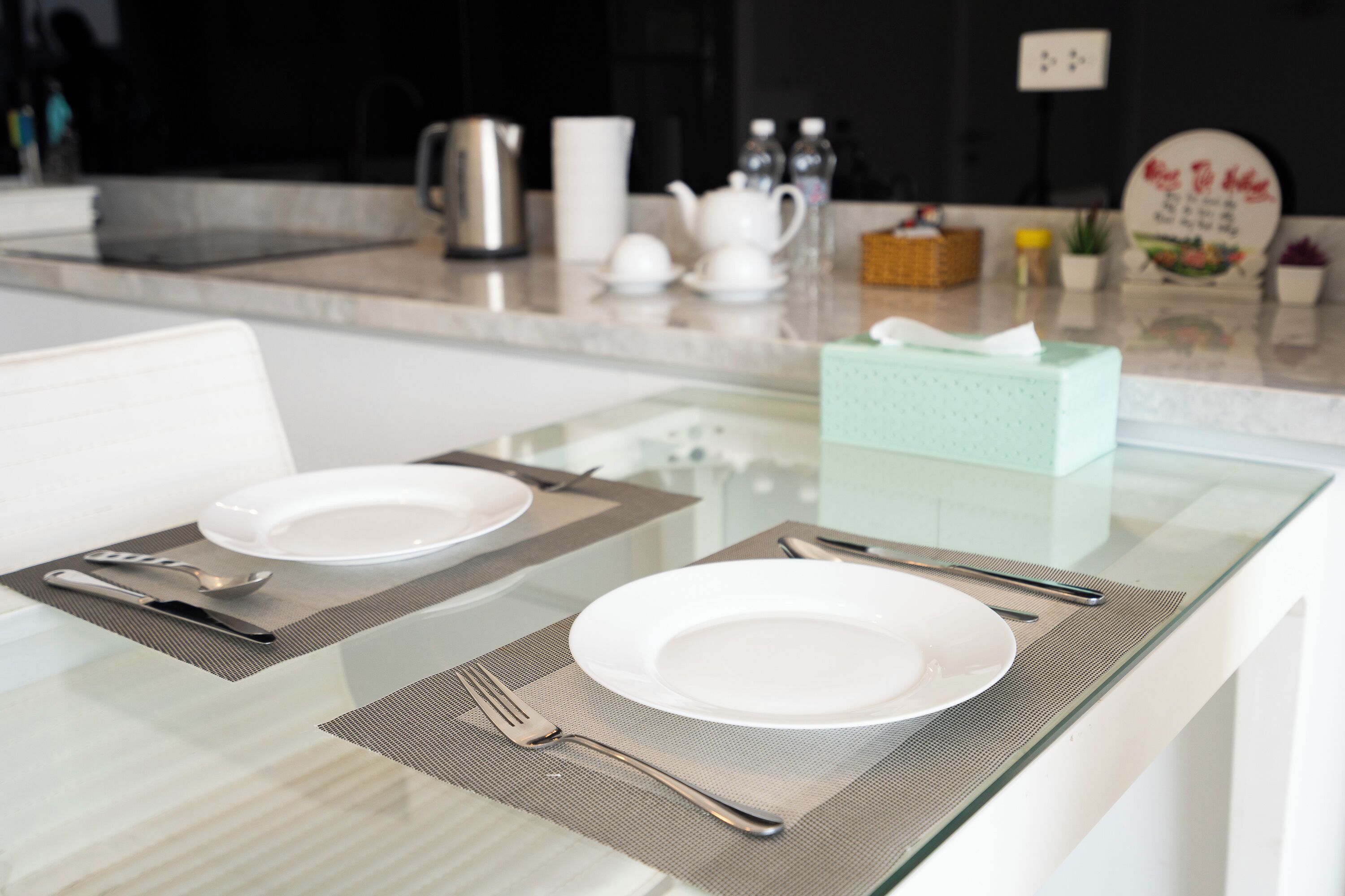 A modern kitchen with a glass dining table set for two, equipped with basic tableware.