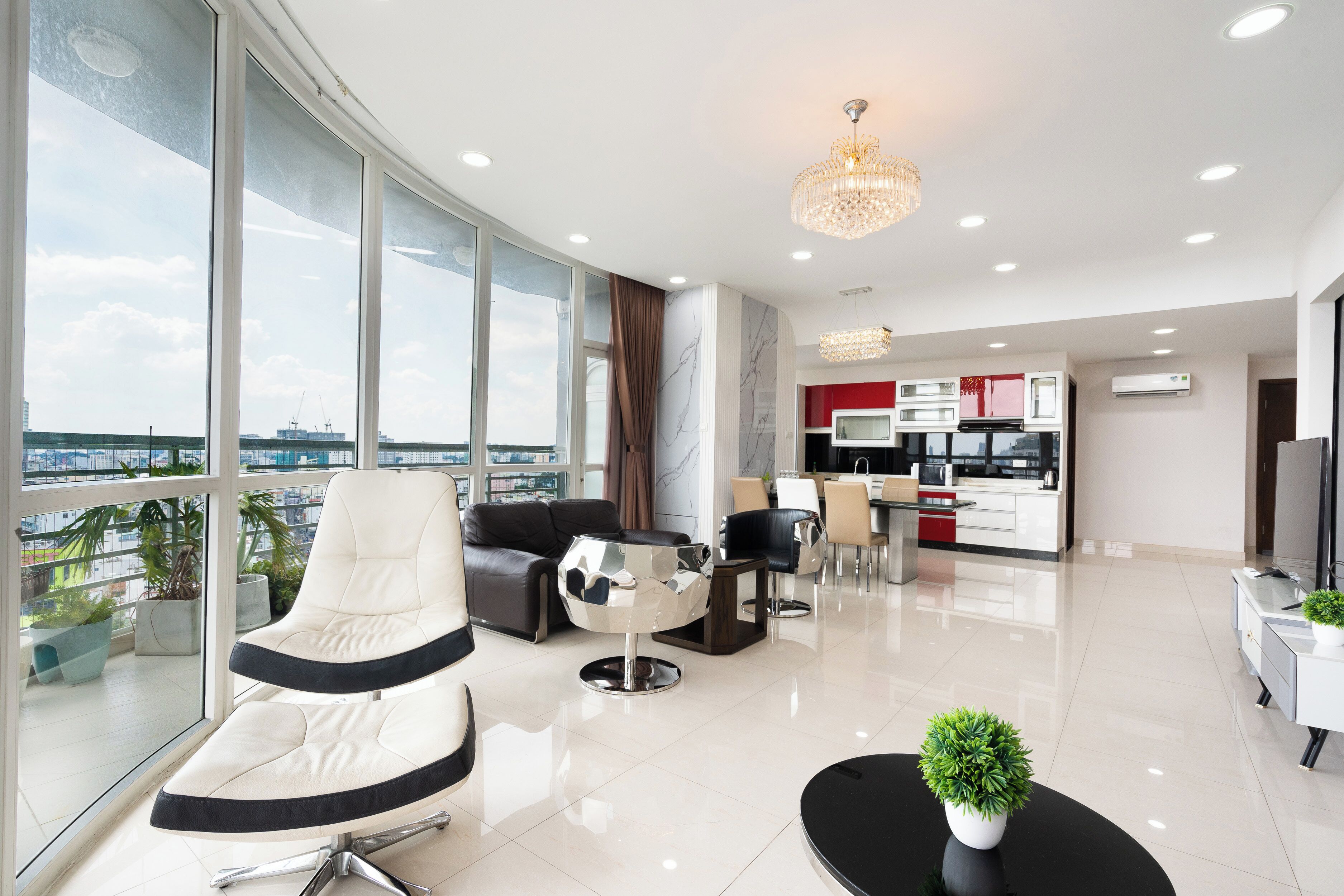 A modern living room with a large window, a sofa, a coffee table, and a dining area with chairs and a kitchen in the background.