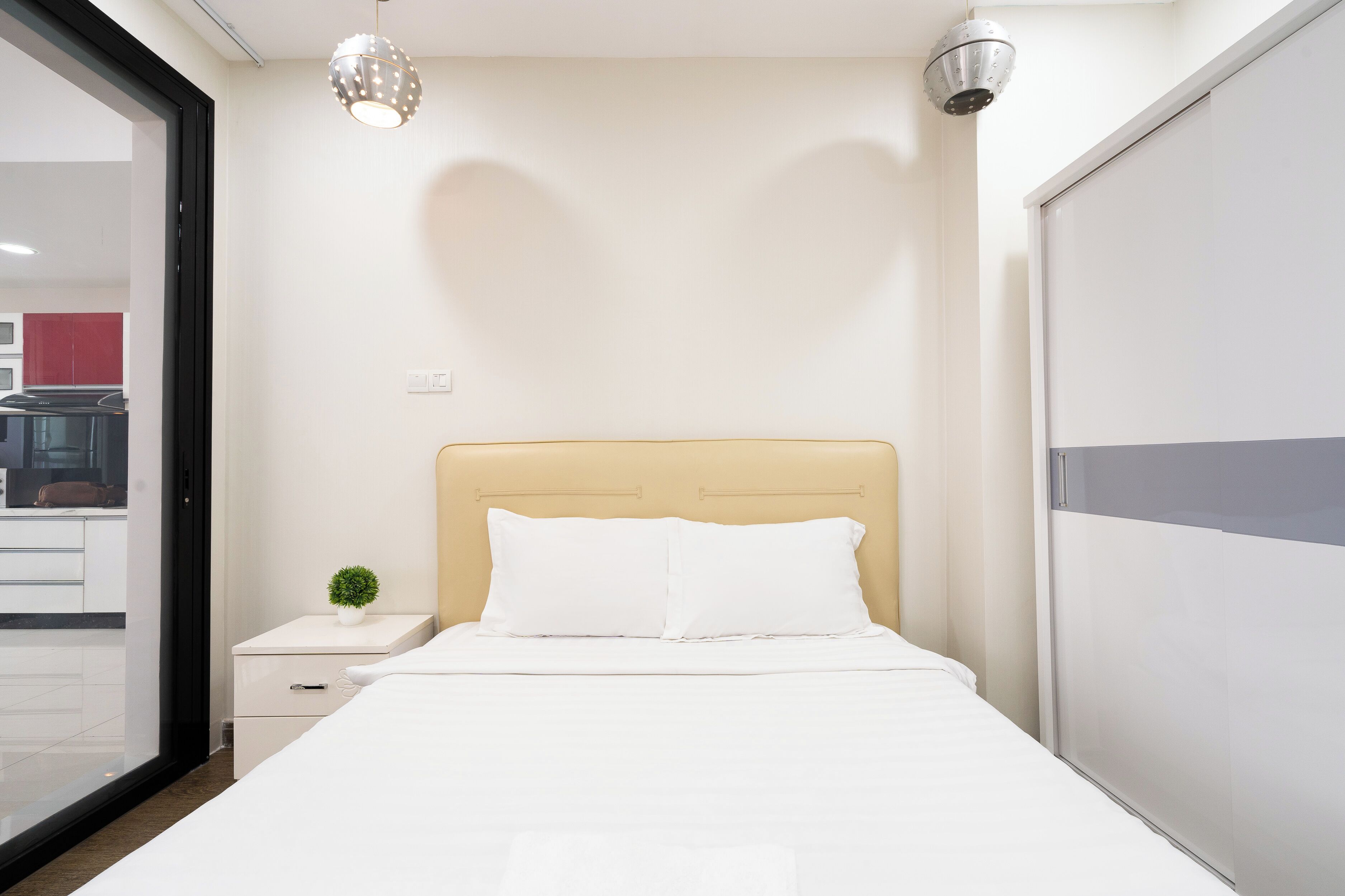 A neatly made bed with white bedding, a small bedside table with a plant, and a modern pendant light.