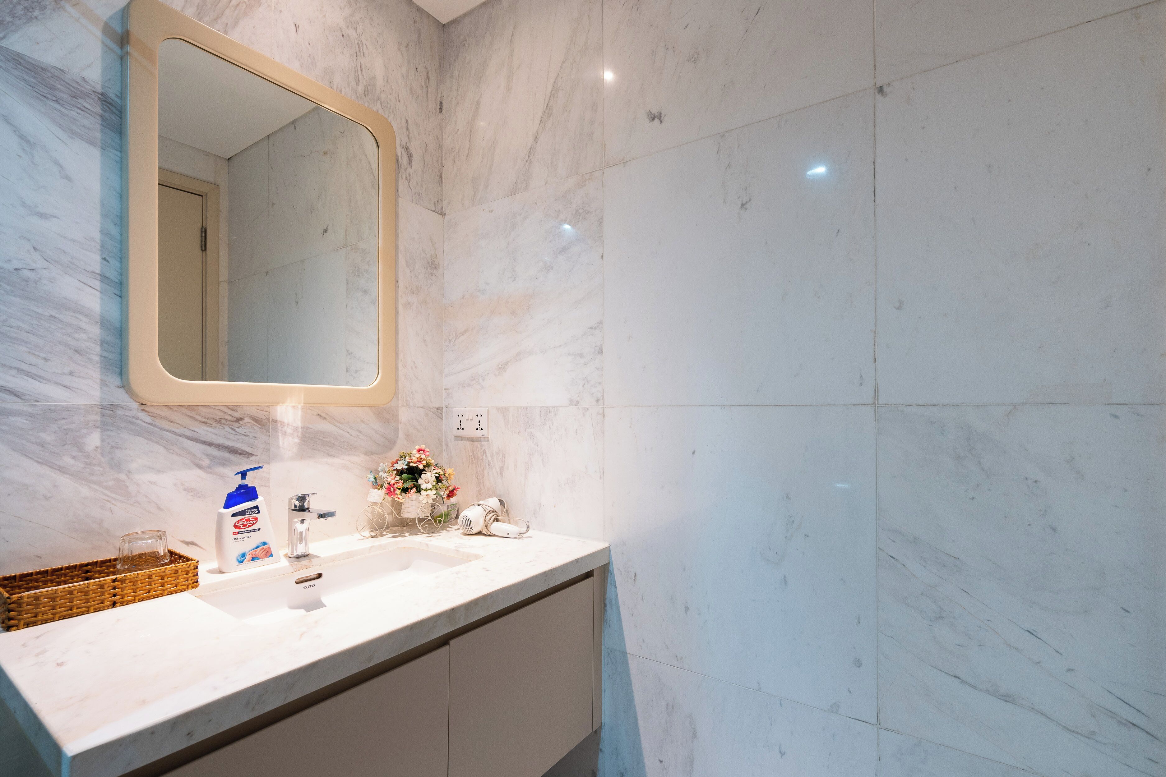 Un baño con pared de mármol, un lavabo con espejo, una botella de jabón para manos y una jarra con flores.
