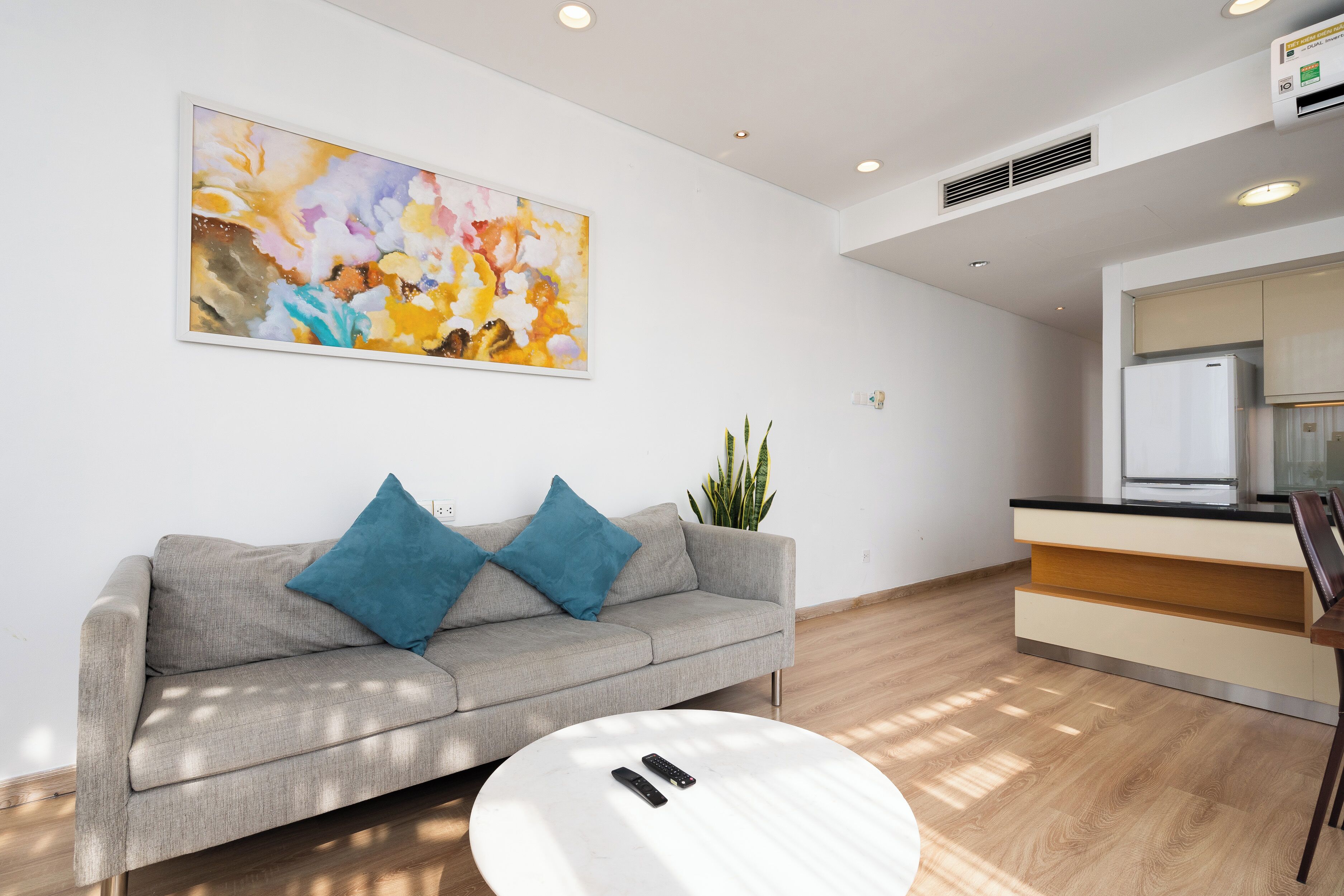 A modern living room with a grey sofa, two blue cushions, a white coffee table, a large abstract painting, a potted plant, and a kitchen area with a refrigerator.