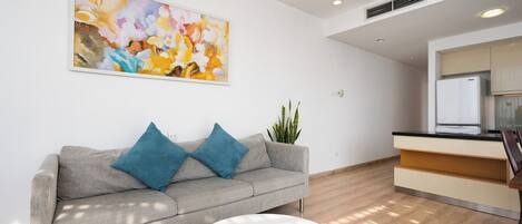 A modern living room with a grey sofa, two blue cushions, a white coffee table, a large abstract painting, a potted plant, and a kitchen area with a refrigerator.