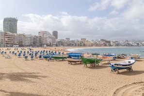 A beach with rows of sun loungers, a row of boats, and a cityscape in the background. - Vacation Apartment 'Casita Sunwaves' with Sea View, Wi-Fi and Air Conditioning (Las Palmas de Gran Canaria)