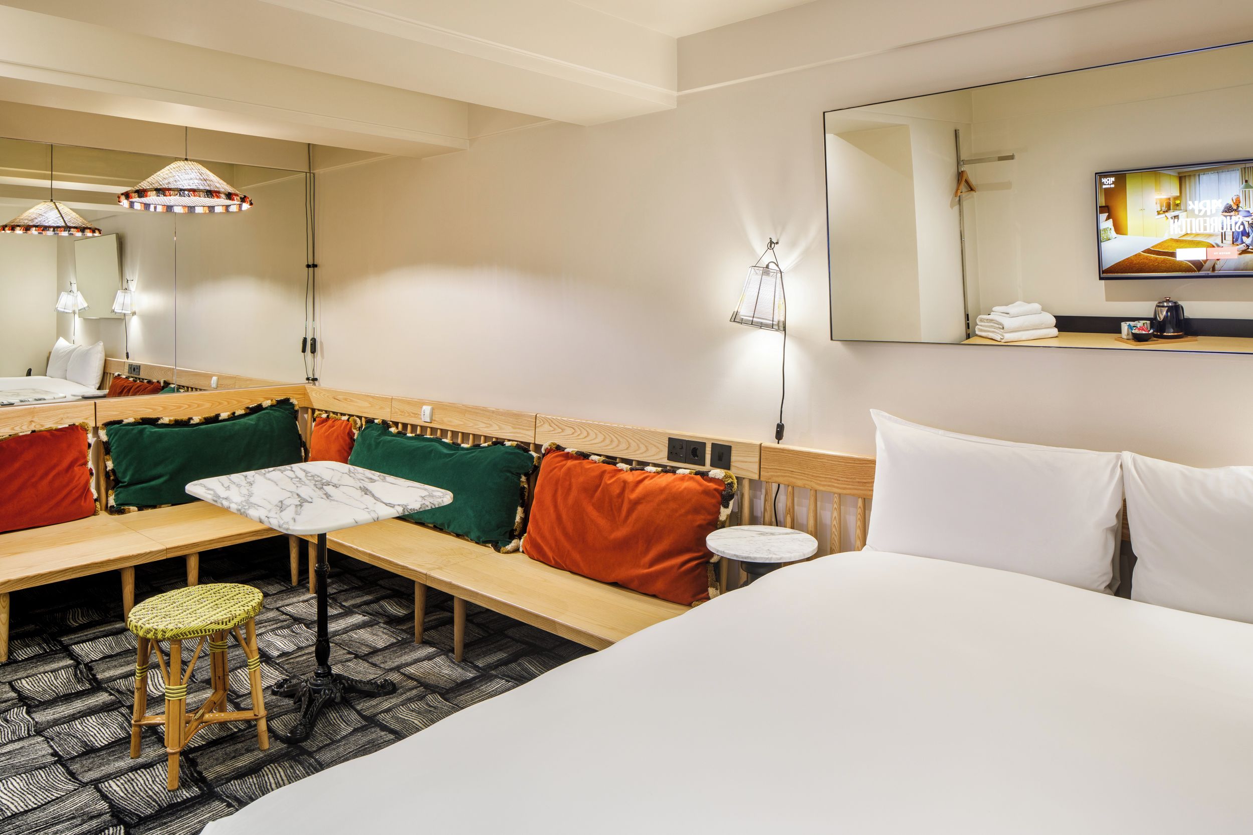 A hotel room with a bed, a small table, a bench, a chair, a mirror, and a television.