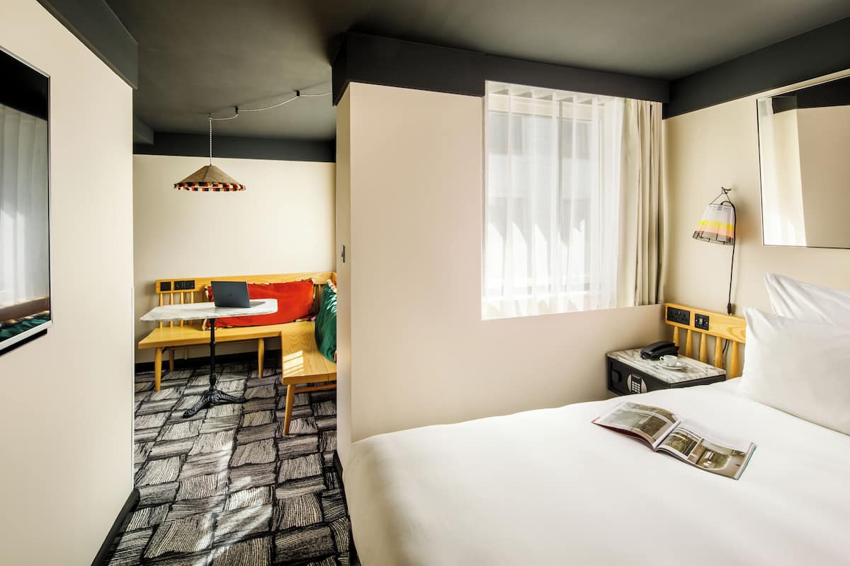 A hotel room with a bed, a desk, a chair, a lamp, a bedside table, and a window.
