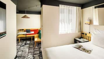A hotel room with a bed, a desk, a chair, a lamp, a bedside table, and a window.