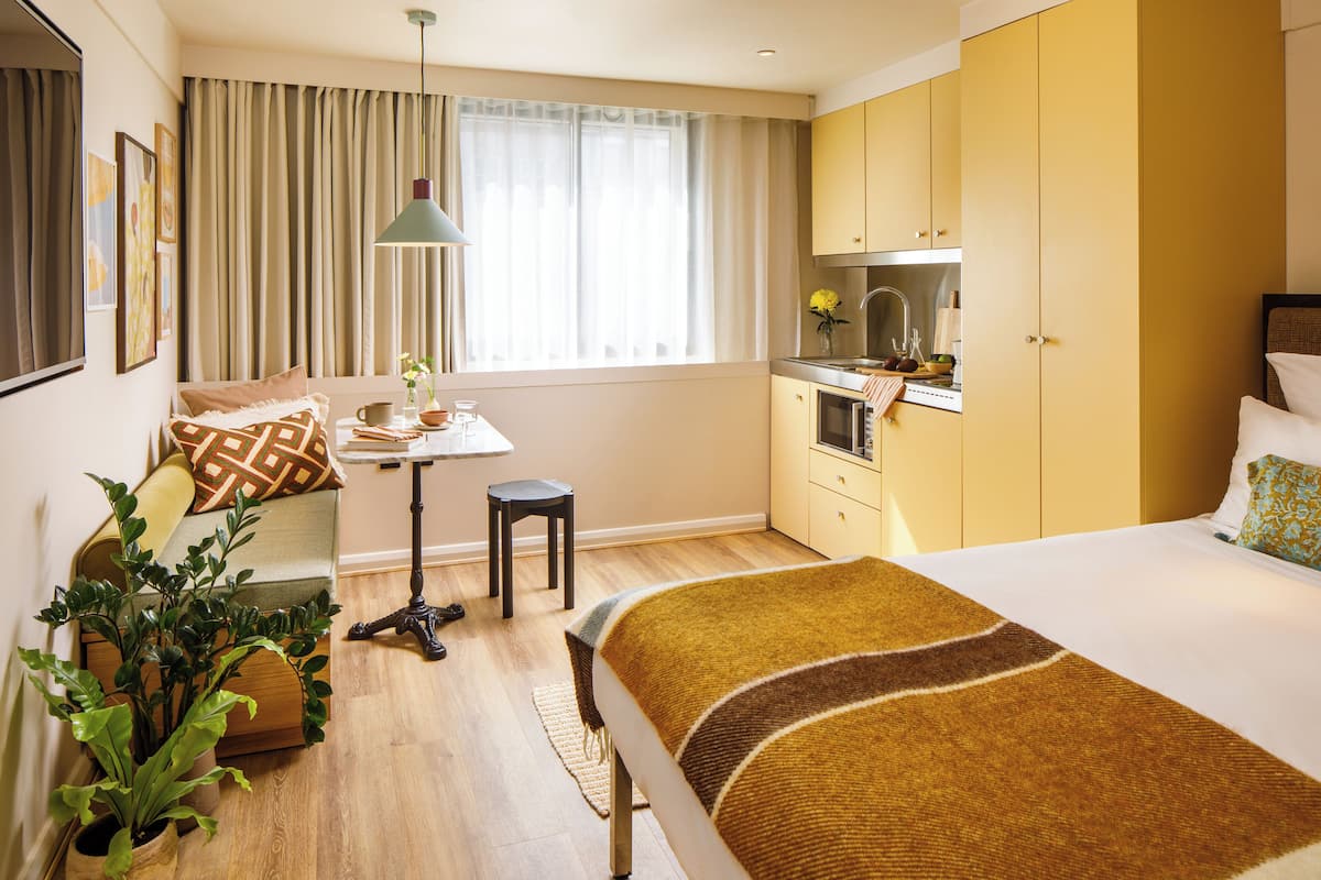 A hotel room with a bed, a desk, a chair, a potted plant, a window with curtains, and a kitchenette.