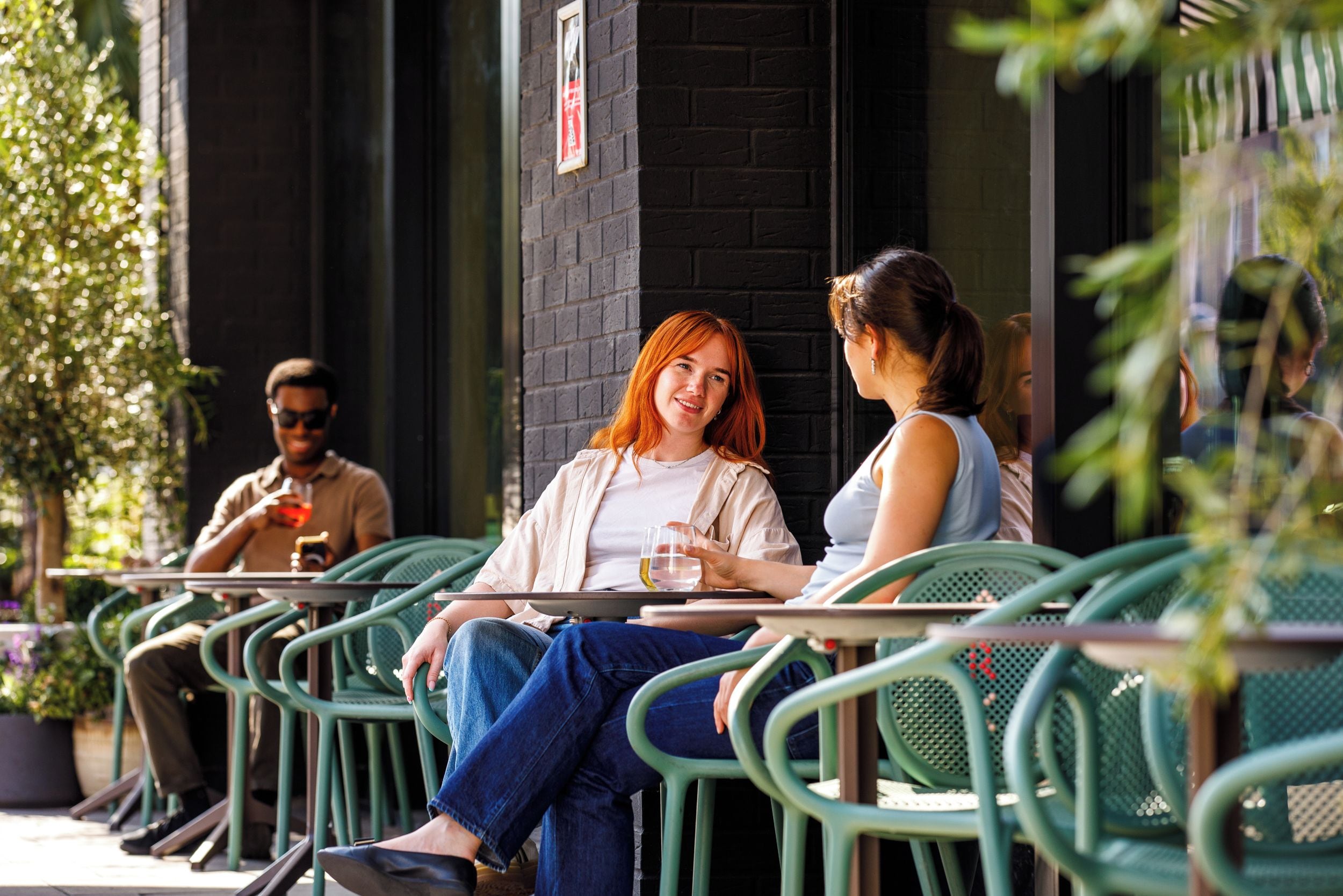 An outdoor seating area with green metal chairs and tables, a brick wall, and a person holding a glass.