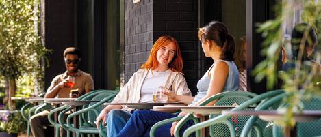An outdoor seating area with green metal chairs and tables, a brick wall, and a person holding a glass.