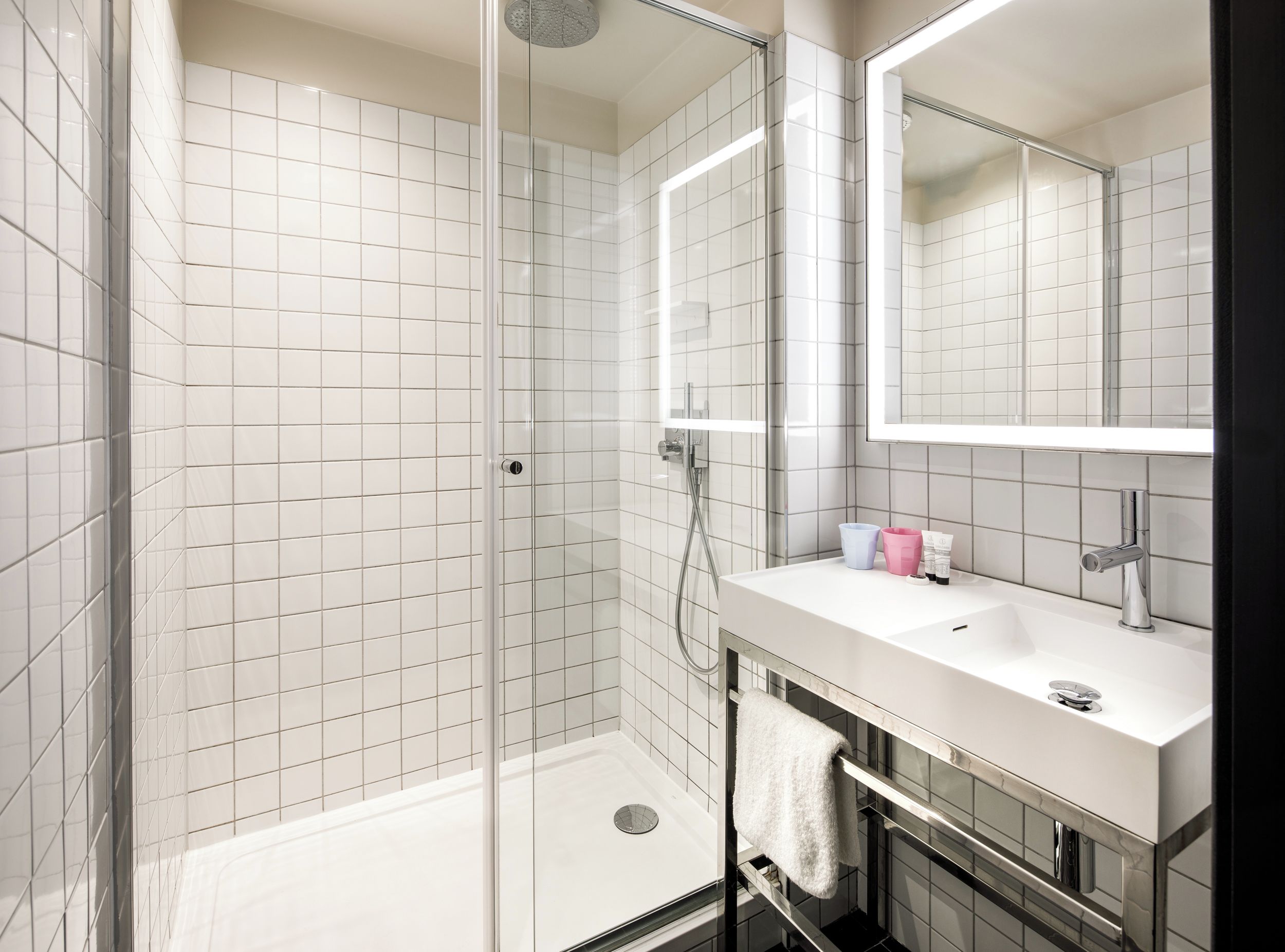 Une salle de bain moderne avec une douche à parois de verre, des murs recouverts de carreaux blancs, un lavabo blanc avec un miroir et un porte-serviettes.