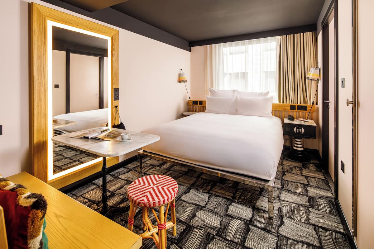 A hotel room with a bed, a desk, a chair, a mirror, a nightstand, and a window with curtains.
