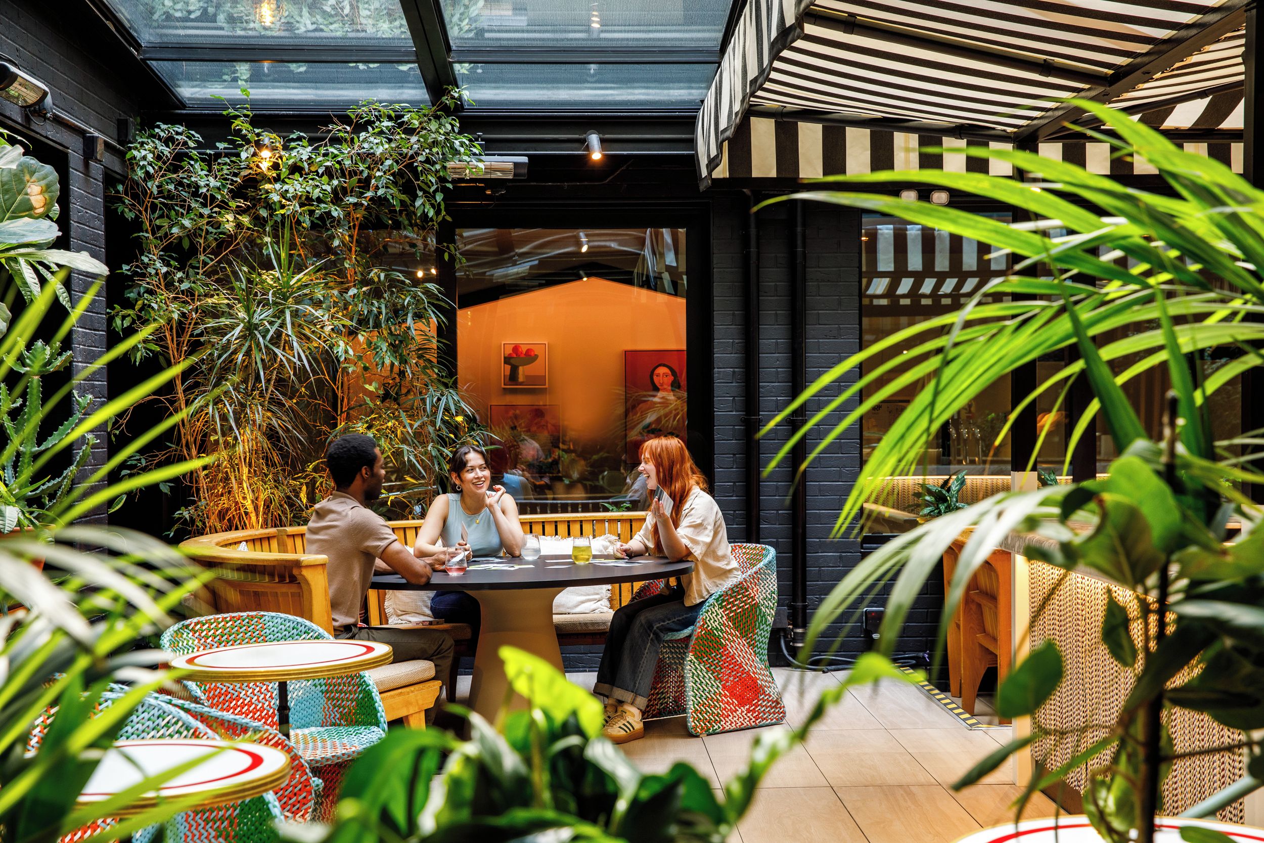 An indoor seating area with round tables and wicker chairs, surrounded by greenery and a glass ceiling.