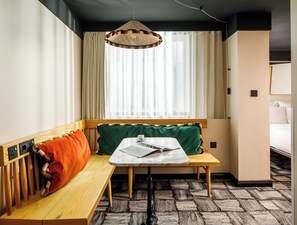 A hotel room with a wooden bench, a small table with a book and a cup, a hanging lamp, and a bed in the background. - ARK Shoreditch (London)