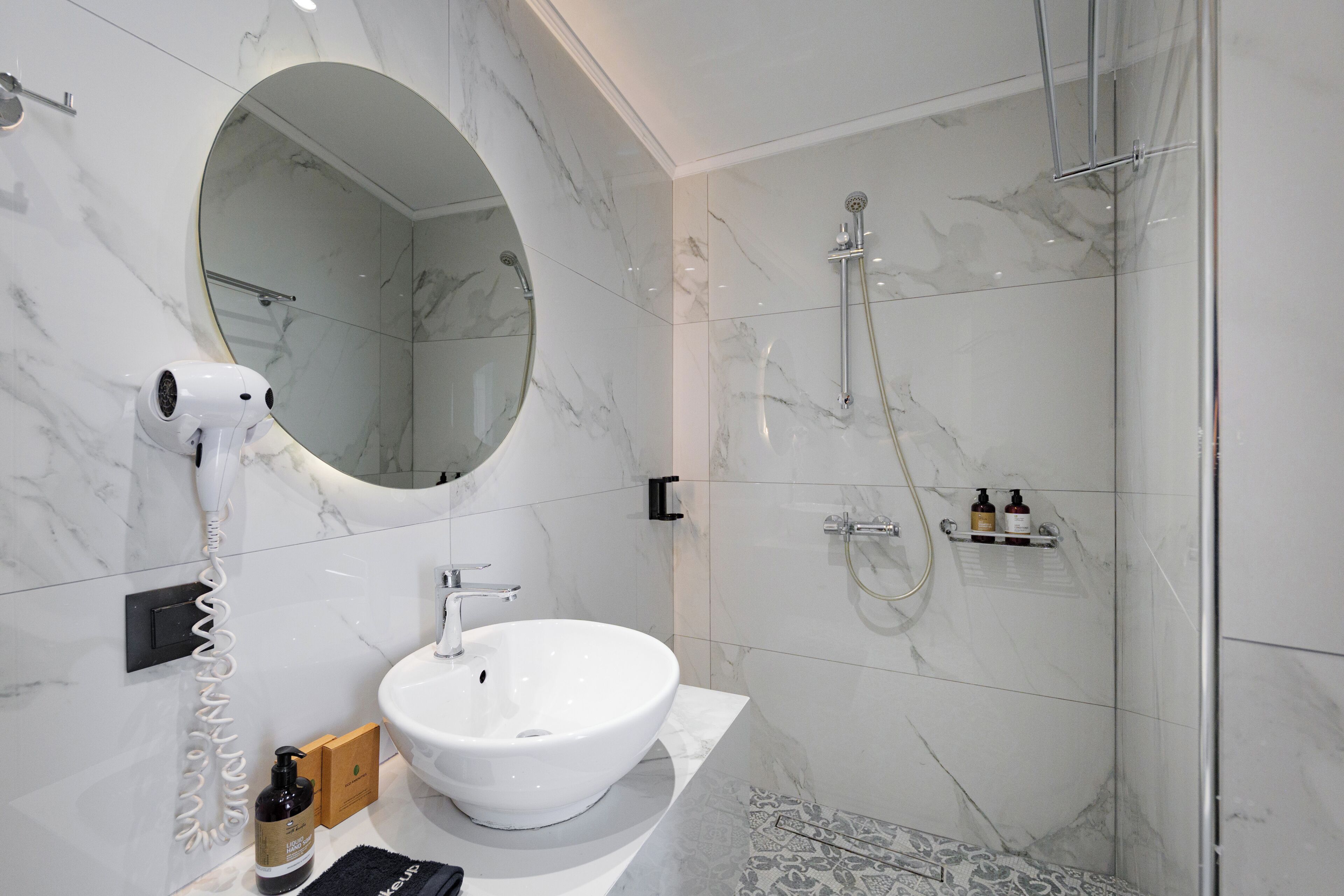A bathroom with a white sink, a round mirror, a handheld hair dryer, and a glass-enclosed shower.