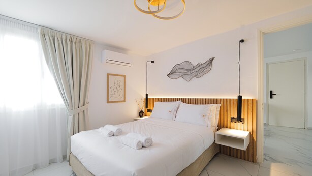 A modern bedroom with a large bed, bedside tables, a wall-mounted light fixture, and a framed artwork. - Kismet Urban Boutique (Heraklion)