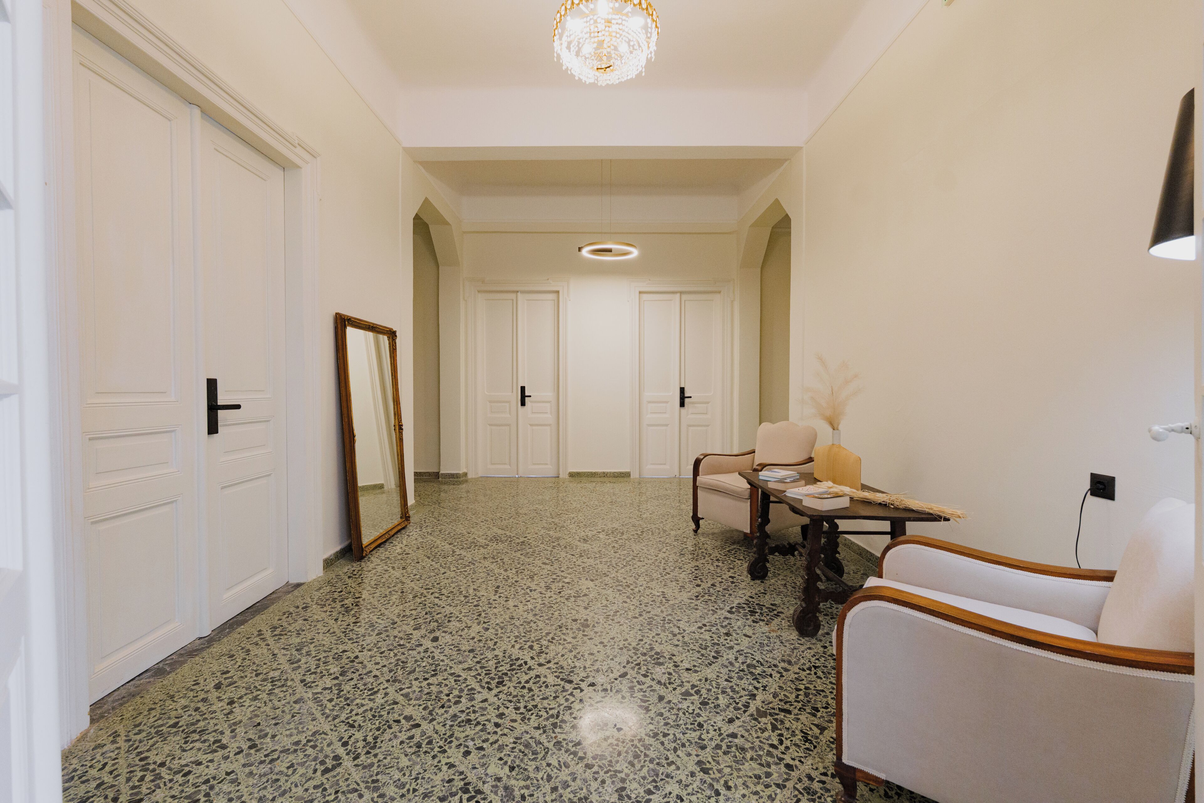 A hallway with a chandelier, a mirror, a table with a lamp, and two chairs.