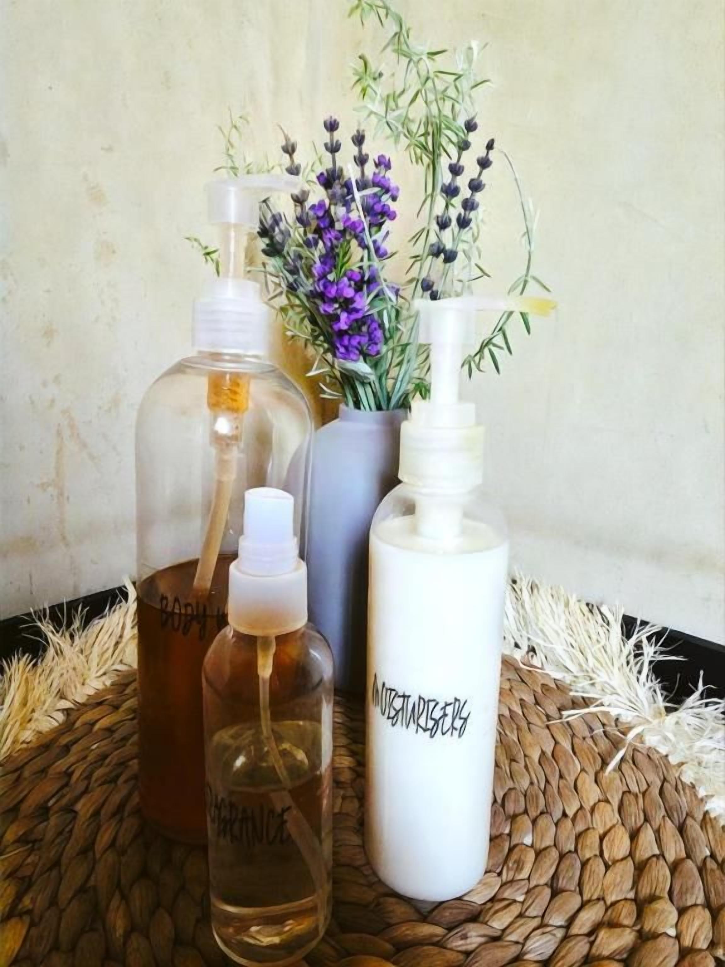 Trois bouteilles de liquide sont posées sur une surface tissée, et un vase rempli de fleurs violettes se trouve à l’arrière-plan.