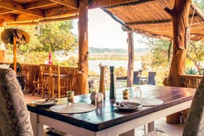 An outdoor dining area with a table set for two, surrounded by wooden chairs, overlooking a body of water. - Frontier River Resort (Vioolsdrif)