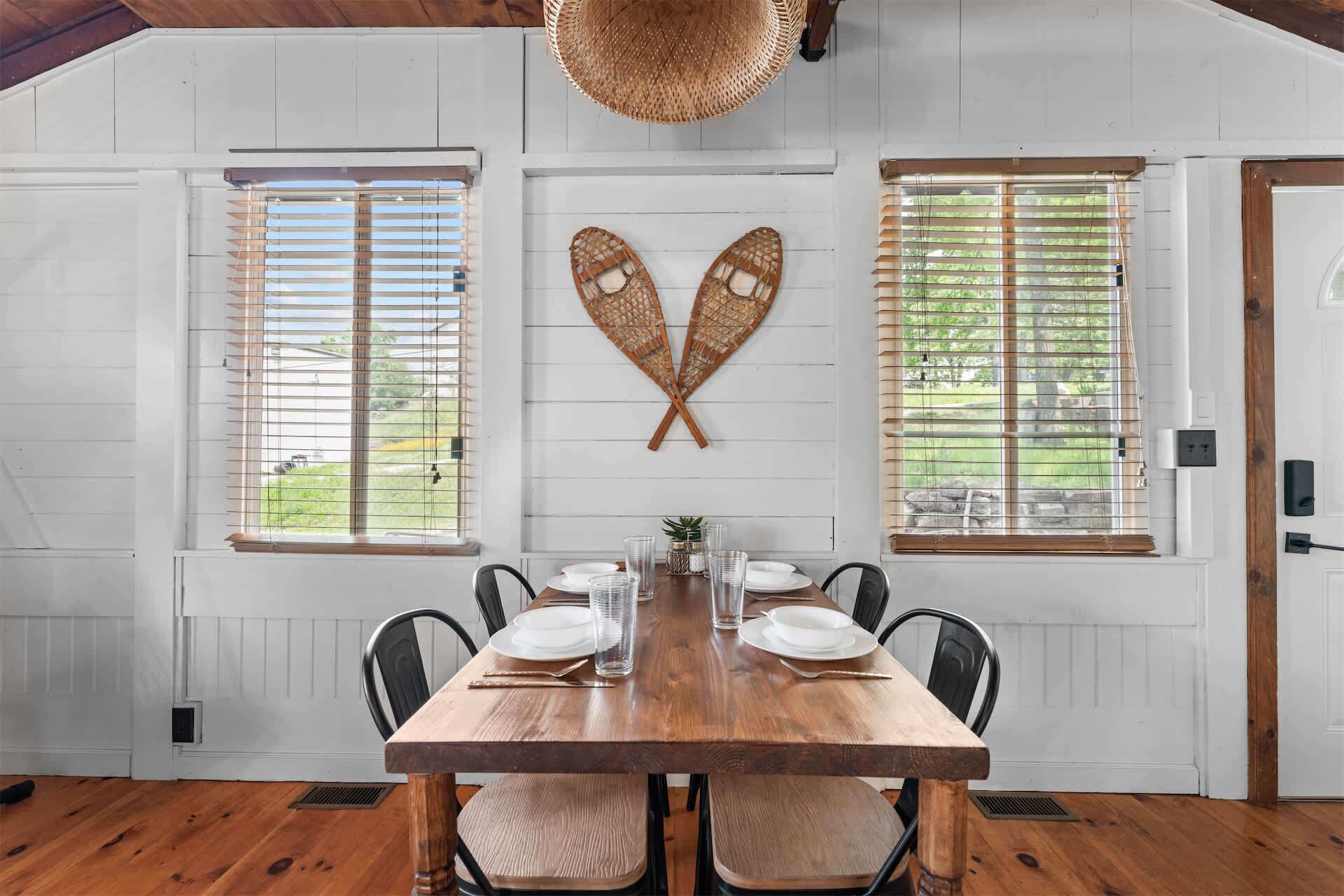 Comedor con una mesa de madera dispuesta para cuatro personas, paredes blancas y dos ventanas con persianas.