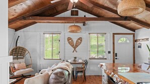 Un interior rústico con vigas de madera a la vista, una zona de comedor con mesa y sillas, un sillón de mimbre y una cocina con encimera de madera.