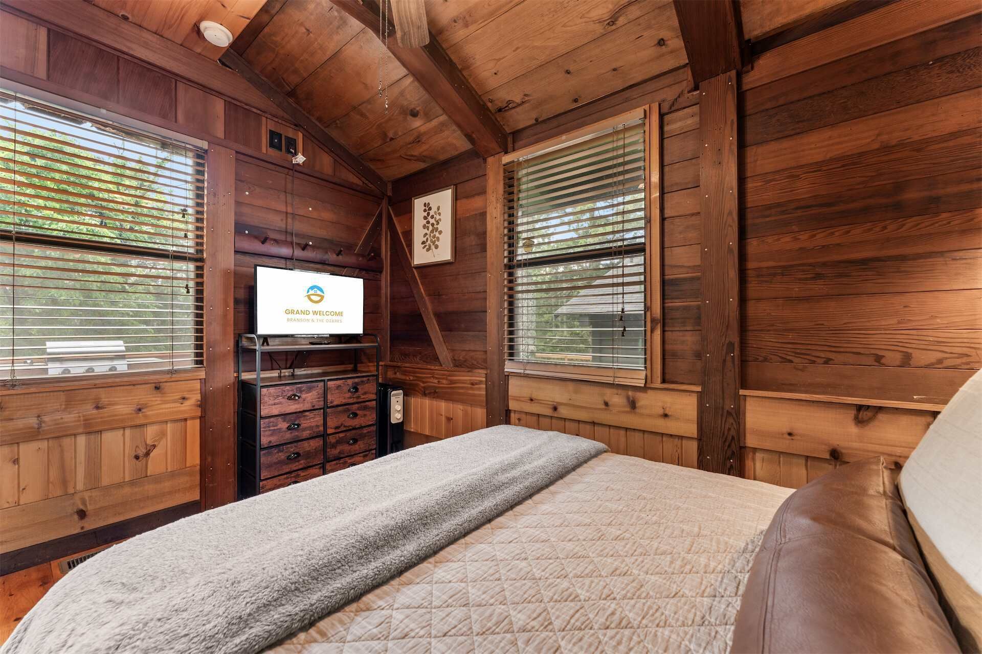 A wooden cabin interior with a bed, a television displaying 'GRAND WELCOME BRANSON & THE CLARKS,' and a fireplace.