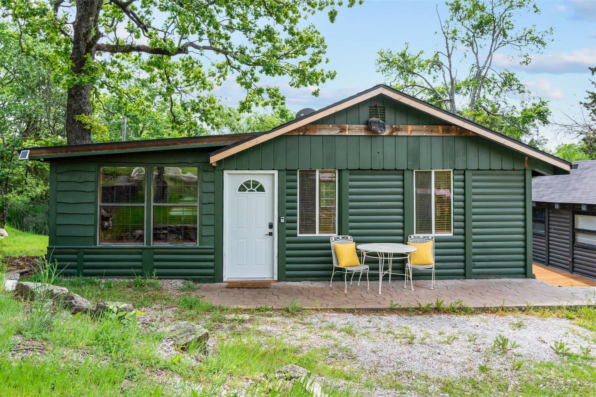 A small cabin with a front door, two windows, a porch with a table and chairs, and a gravel path.