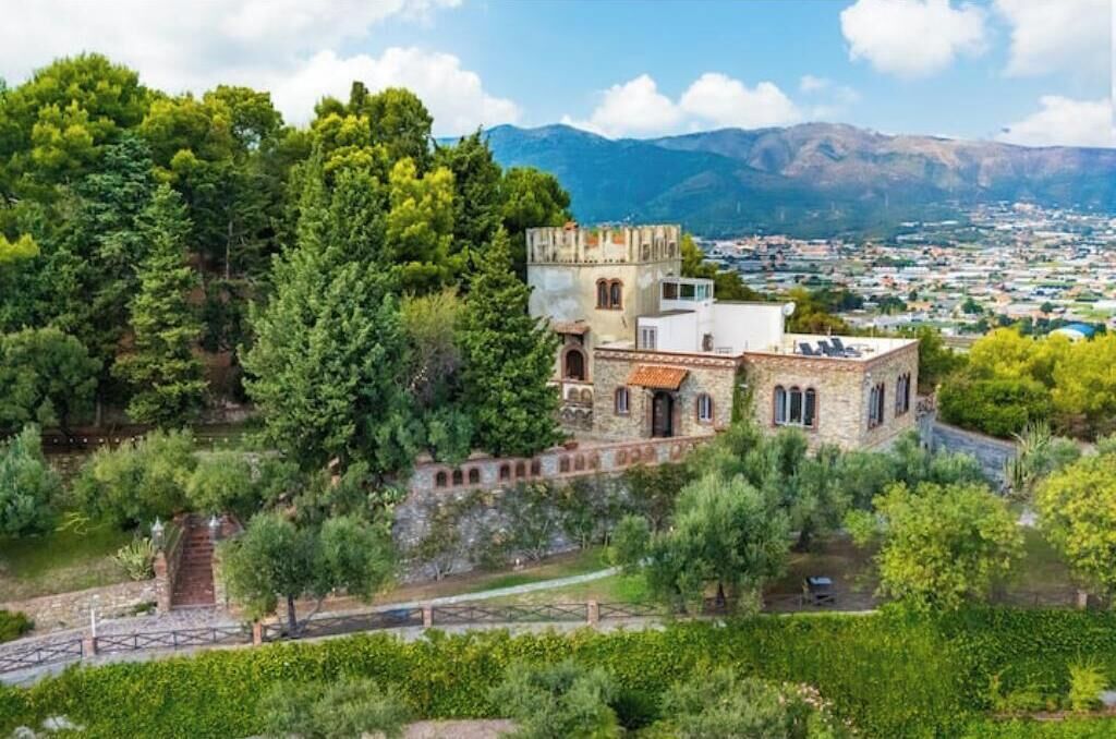 A historic stone building with a tower, surrounded by lush greenery and a mountainous backdrop.