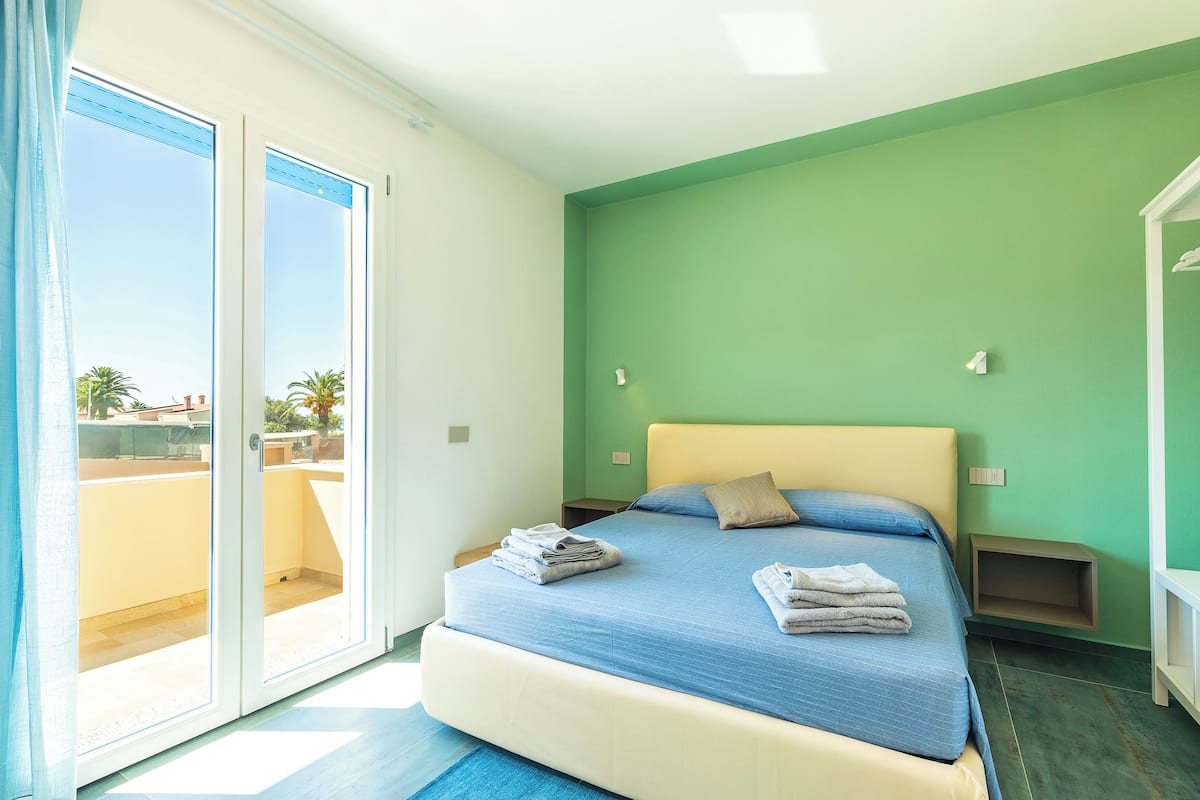 Une chambre à coucher comprenant un lit, une table de chevet, une étagère fixée au mur et une porte-fenêtre coulissante donnant sur un espace extérieur.