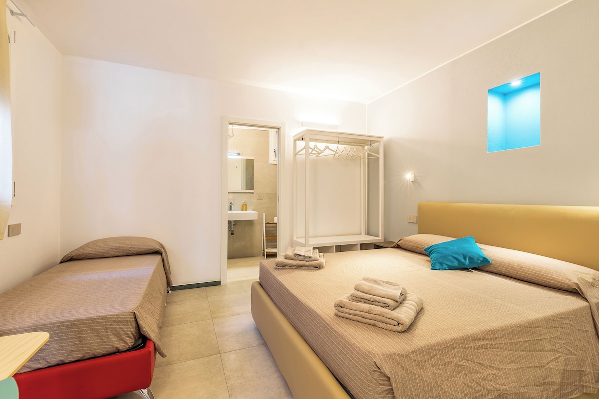 Une chambre d’hôtel avec un lit, une chaise rouge, une armoire et une salle de bain visible à travers une porte ouverte.