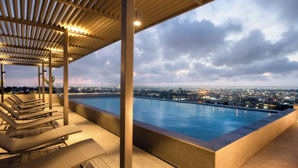 A rooftop pool with lounge chairs, a view of the cityscape, and a covered area with lighting. - The Lennox by Clifton Homes (Accra)