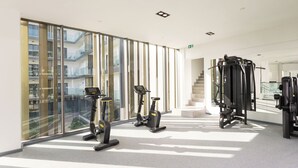 A modern gym with exercise equipment, large windows, and a view of a building. - The Lennox by Clifton Homes (Accra)