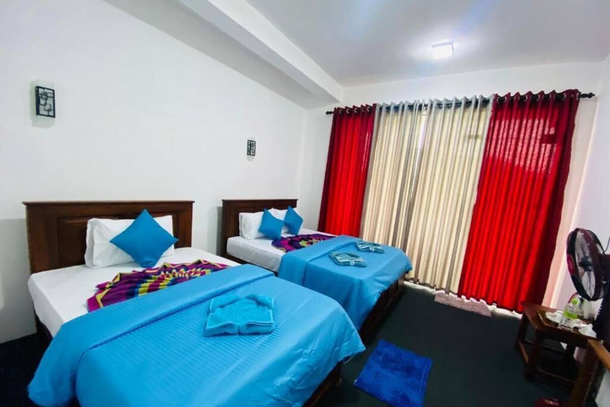 Habitación de hotel con dos camas, un escritorio, una silla, un ventilador y una ventana con cortinas rojas.