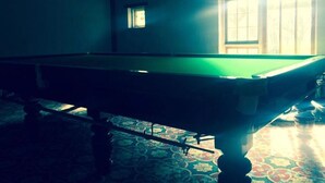 Un tavolo da biliardo in una stanza con tappeto a motivi, una piccola finestra e una ventola a soffitto.
