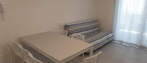 A room with a white table and four chairs, a striped sofa, a telephone on the wall, and a window with sheer curtains.