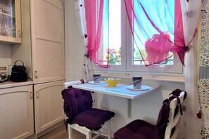 Una cocina con una mesa blanca, dos sillas moradas, un bol amarillo y una cafetera.
