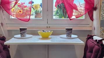 A table with two coffee cups and a yellow bowl, decorated with red bows, in front of a window with potted plants.