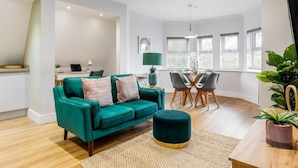 A modern living room with a teal sofa, a round ottoman, a dining area with a table and chairs, and a large window with blinds.