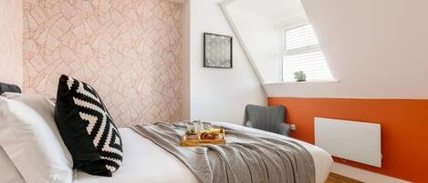 Slaapkamer met een bed, kussens, een dienblad met eten, een stoel, een radiator en een raam met jaloezieën.