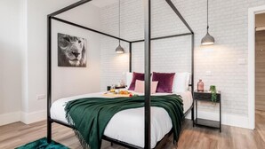 A four-poster bed with a green throw, a nightstand with a plant, and a framed picture on the wall.
