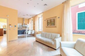 A spacious living area with a sofa, a dining table, and a kitchen in the background. - Hostdomus - The Mistral Apartment (Alassio)