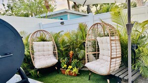 Two wicker chairs with cushions, a barbecue grill, and a small potted plant with yellow flowers.
