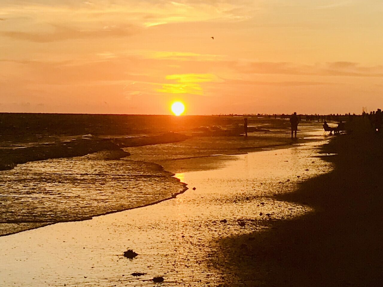 Ein Sonnenuntergang über einem Strand mit ruhiger See und einigen Menschen, die am Ufer entlanggehen.