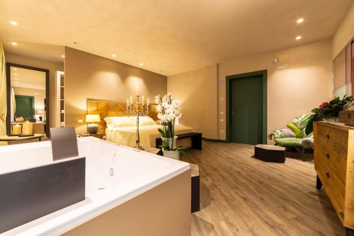 A modern hotel room with a large bed, a sitting area with a sofa, a wooden dresser, and a bathroom with a bathtub.