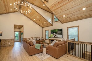 Ein geräumiges Wohnzimmer mit hoher Decke, Holzbalken, einem Kamin und einem großen Fenster.