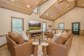Ein großzügiges Wohnzimmer mit hoher Decke, Holzbalken, einem Kamin, Ledersofas und einem großen Fernseher.